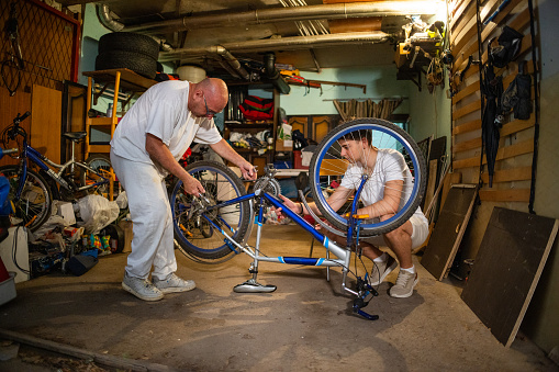 Dedicated Caucasian father and teenage son together repair bicycle at the messy garage