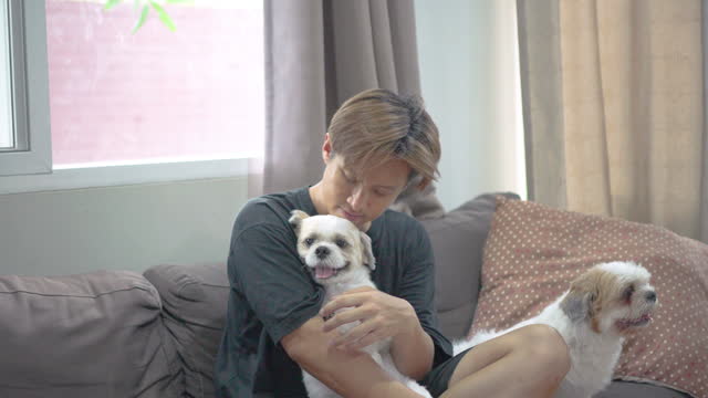 Asian man embracing and petting his dog while sitting on a sofa in the living room at home.