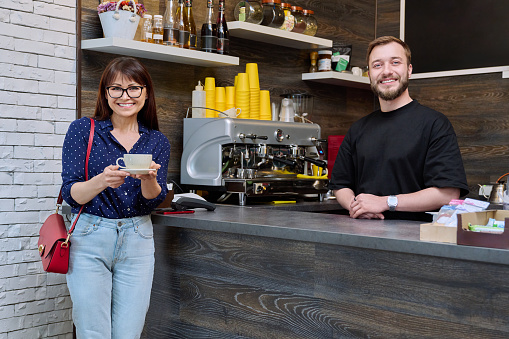 Young male barista talking to woman customer in coffee shop, near counter with cup of freshly prepared cappuccino coffee. Small business, service, coffee shop, staff, work concept