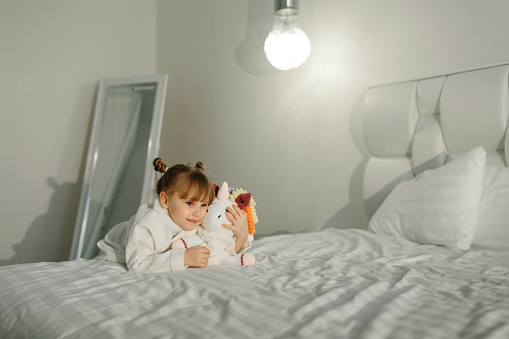 A girl in a bathrobe plays on the bed with a knitted unicorn