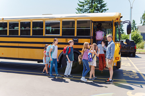 A male teacher or bus driver stands by the open door of a school bus and greets with a high five a multiethnic group of elementary age students as they board the bus.