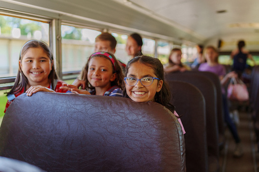 A multiracial group of elementary age girls sit together on a school bus and smile directly at the camera.