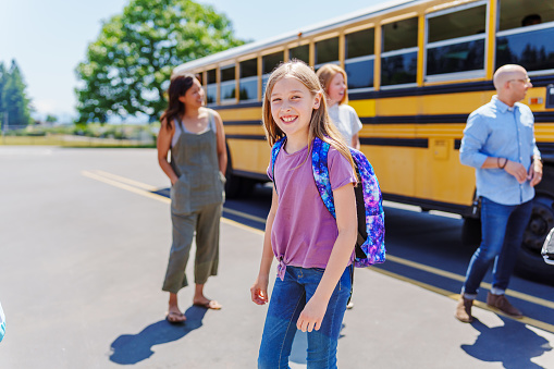 An elementary age girl smiles confidently at the camera as she walks away from her school bus after arriving at her bus stop on a warm and sunny day. Parents waiting to greet their children are standing in the background.