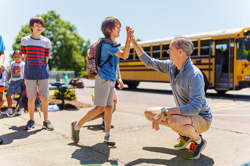 A kind male teacher squats down to greet elementary age kids with a high five in front of the school on a warm and sunny day. A parked school bus is visible in the background.