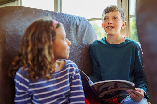 A tween boy sits on a school bus with his younger sister and holds a book open while talking cheerfully with a friend who is out of frame. The boy's sister is looking at him and listening to what he has to say.