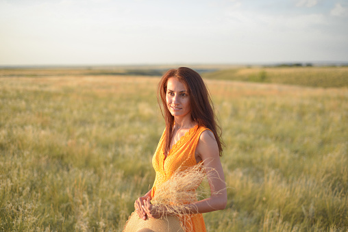 A pretty young girl with long brown hair in an orange dress in a field at sunset. The concept of freedom and nature.