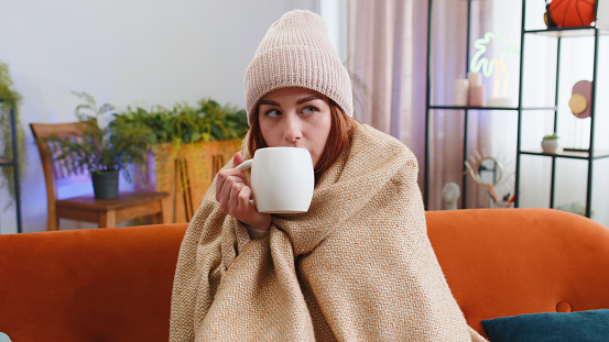 Sick redhead woman wear hat wrapped in plaid sit alone shivering from cold on couch drinking hot tea in unheated apartment without heating due debt. Unhealthy girl feeling discomfort try to warming up