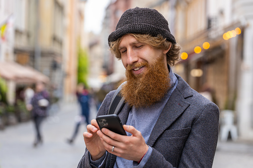 Portrait of young adult man using smartphone typing text messages looking for a way on map in mobile navigator app outdoor. Redhead bearded guy walking in urban city street background. Town lifestyles