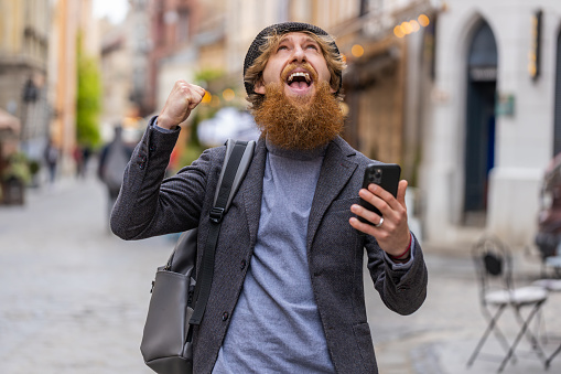 Young bearded man use mobile smartphone celebrating win good message news, lottery jackpot victory, giveaway online. Happy redhead guy walking outdoors in urban city street background. Lifestyles