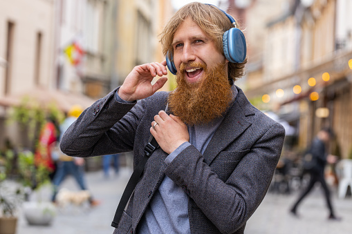 Happy relaxed overjoyed young bearded man in wireless headphones choosing, listening favorite energetic disco rock n roll music in smartphone dancing outdoors. Tourist guy walking in urban city street