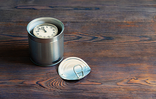 An old clock lies in an open tin can. canned time. There are dark wooden boards of the table and a removed cover. Background. Concept.