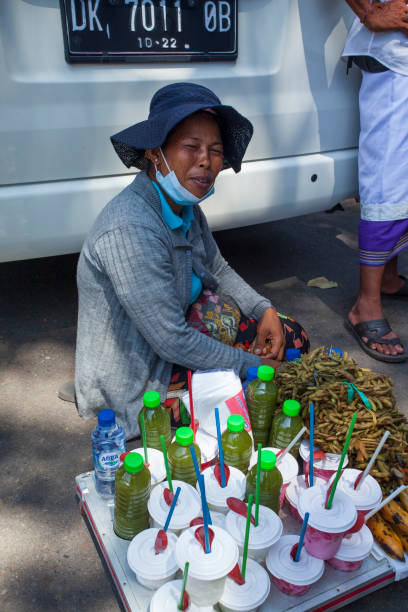 una mujer balinesa local sentada en el suelo y vendiendo bebidas tradicionales en un día muy caluroso fuera del templo pura goa lawah en klungkung, east bali, indonesia. - pura goa lawah fotografías e imágenes de stock