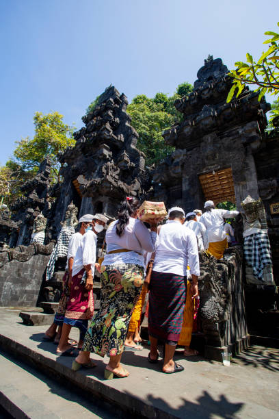 cerimonia balinese a pura goa lawah o tempio di goa lawah a east bali con persone che indossano abiti tradizionali, kebaya, parei batik e che portano offerte e con uomini che indossano cappelli udeng ed entrano nel tempio attraverso gli ingressi principali - pura goa lawah foto e immagini stock