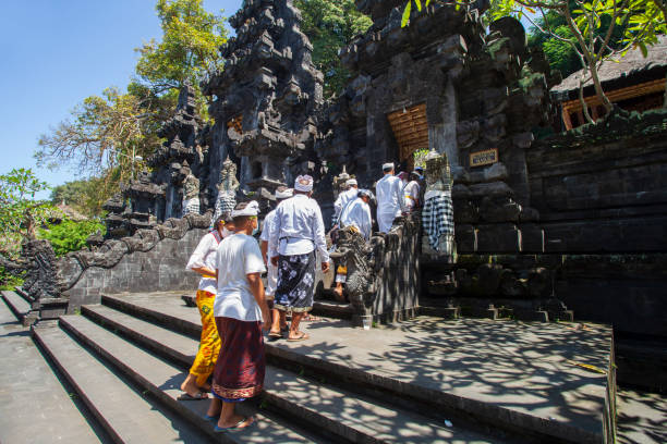 ceremonia balinesa en pura goa lawah o templo de goa lawah en el este de bali con personas vestidas con trajes tradicionales, kebaya, pareos batik y llevando ofrendas y con hombres con sombreros udeng y entrando en el templo a través de las entradas princ - pura goa lawah fotografías e imágenes de stock