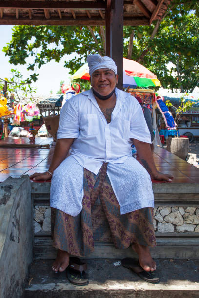 un homme balinais souriant vêtu d’une robe traditionnelle avec une chemise blanche, un batik saring et un udeng, à l’extérieur du temple pura goa lawah ou du temple bat cave à goa lawah, klungkung regency, east bali lors d’une cérémonie hindoue - pura goa lawah photos et images de collection