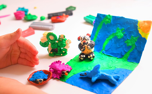 Child smearing colorful plasticine on cardboard and creating fairy tale card with cartoon animals, panda, birds, shark. By spreading, modelling and adding texture to plasticine child creating