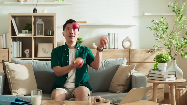 Smiling Attractive handsome Asian male adult stay home on sofa relax playful fun practice juggling with fresh fruit apple at home