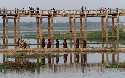 Mandalay, Myanmar - 3/8/2020 : Groups of people walk on U Bein Bridge as a group of buddhist monks walk on footpath