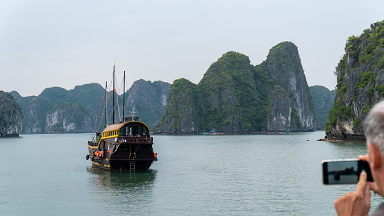 Halong Bay, Vietnam - 11/19/2019 : Man taking a picture with cell phone of a junk boat.