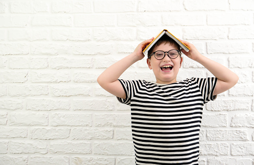 A cute cheerful boy in stripped tee shirt and glasses and hold a yellow book on his head on white bricks background.