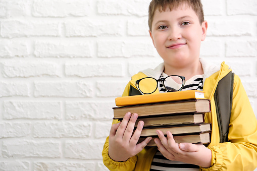 A cute cheerful boy in stripped tee shirt, yellow jacket, holds books on white bricks background.