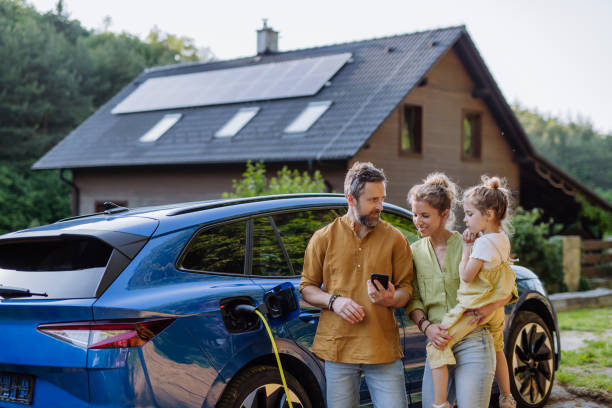 Family with little girl standing in front of their house with solar panels on the roof, having electric car. stock photo