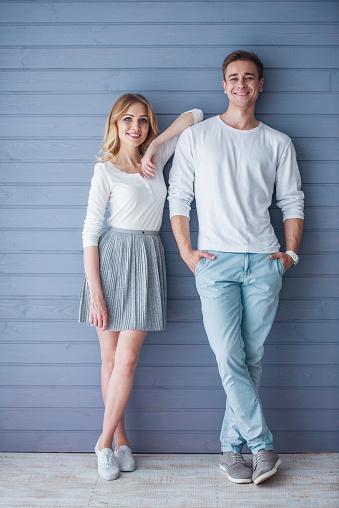 Full length portrait of beautiful young couple in casual clothes looking at camera and smiling, standing against gray wall