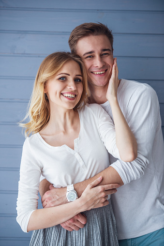 Portrait of beautiful young couple in casual clothes hugging, looking at camera and smiling, standing against gray wall