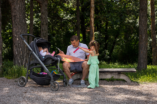 The man is sitting on the bench in the park with his daughters. The baby is in the stroller, and the older daughter is standing next to her dad, and helping him to make the kite so she can fly it later.