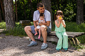 The father sitting on the bench with the baby girl in his hands , watching the older daughter eating the apple.