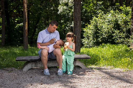 The father is feeding, and holding the baby in his lap. The little girl is sitting on the bench by her father and sister, and eating the yellow apple.