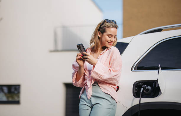 Young woman with smartphone charging her electric car in home, sustainable and economic transportation concept. stock photo