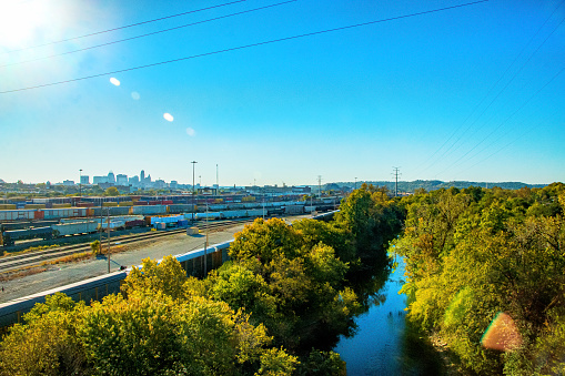 A high vantage view point of a large scale commercial inland cargo container and railroad yard in Cincinnati Ohio.  This is a multi-track freight train transfer station.  There is a river flowing beside this complex and it is surrounded by lush trees. The river is Mill Creek. There is naturally occurring lens flare from the morning sun that burst on the left of the image.