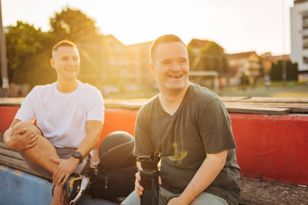 A man with a chromosomal disorder known as down syndrome holds a reusable bottle in his hands while sitting on a bench and having fun with his male friend
