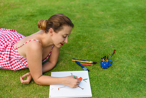 Young woman artist and illustrator drawing witha pencil, laying on a lawn in summer