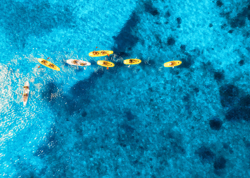 Aerial view of yellow kayaks in blue sea at summer sunny day. People on floating canoes in clear azure water. Sardinia island, Italy. Tropical landscape. Sup boards. Active travel. Top view from drone
