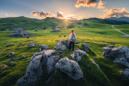 Girl with packpack on the stone, cows on green meadows in beautiful old alpine village in mountains at sunset in summer. Landscape with young woman, houses, trees, sky. Travel and Hiking. Slovenia