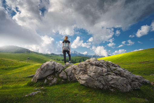 Girl with packpack on the stone, hill with green grass in beautiful alpine mountain valley at sunset in summer. Landscape with young woman in alps, trees, sky with clouds. Travel and Hiking. Slovenia
