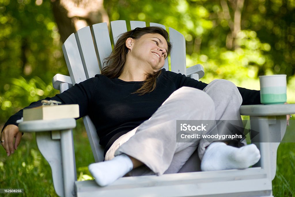 Woman sitting in a chair with coffee cup and book A woman sitting comfortably in an outdoor adirondack chair, with a coffee mug on one arm, and a book on the other. 30-34 Years Stock Photo