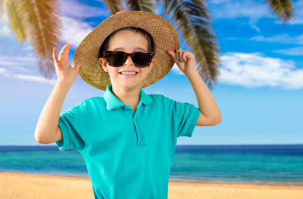 Child on vacation wearing blue t-shirt hat sunglasses at tropical beach happy face smiling and looking at the camera. Positive person
