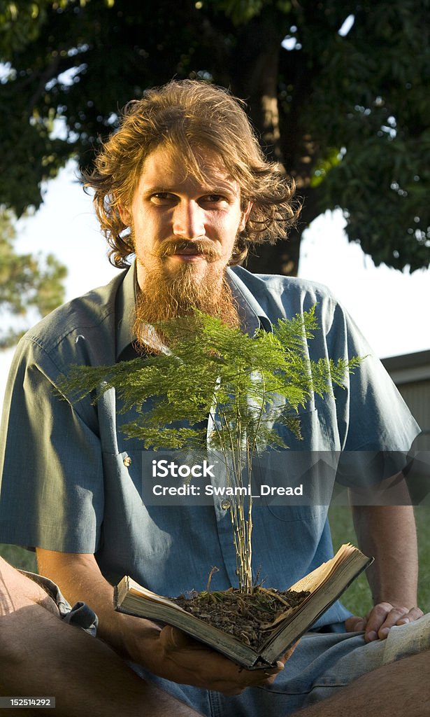 Man reading book with tree growing out of it. Man with long beard sitting outside in nature with a small tree growing out of the book he is reading. Achievement Stock Photo