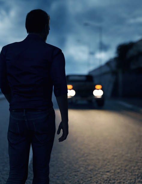istock Man on road walks towards car with illuminated headlights under a stormy sky. 3D render. 1525143766