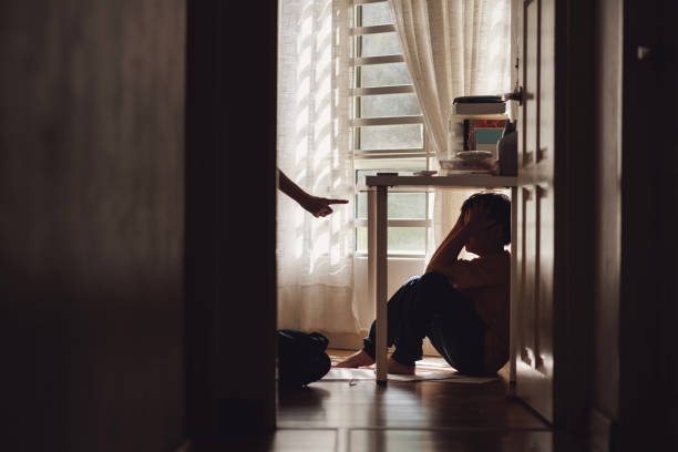 A little Asian boy with a sad and lonely expression hides in the shadows under a table at home, while being scolded by his mother