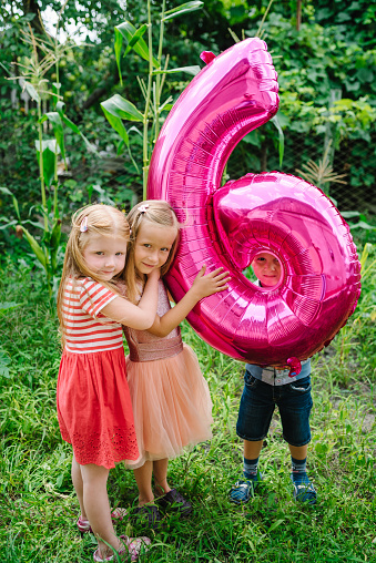 Happy children friends hugging and holding a balloon on sixth birthday in the backyard. Funny emotional kids with pink balloon in the garden. 6 six years old, 6th birthday party celebration.
