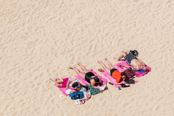 A lonely man sunbathing, wearing a protective face mask and sunglasses sits next to an  empty deck chair, near the parasol and rubber duck inflatable,he is on a sandy beach with the waves crashing behind him
