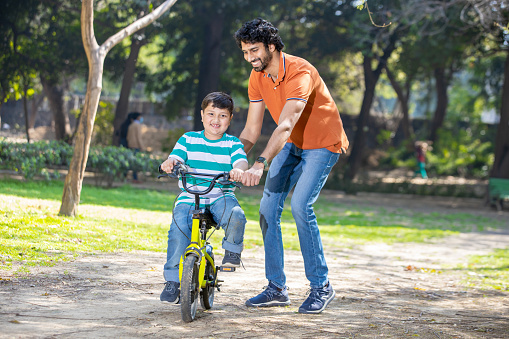 Indian father helping her son learn to ride a bicycle in the park outdoor. kid having fun with bike while his father helping him balance. hobby and activity.