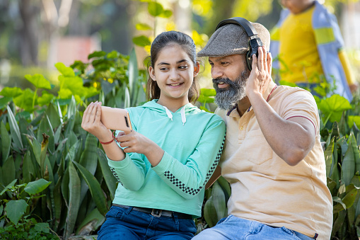 Happy Indian father and daughter using smart phone at park, mature senior man wearing headphone and girl holding smart phone, watching video social media on mobile phone outdoor.