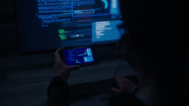 Young man hacker watching smartphone and LCD monitor to hacking data servers from multiple computers in a dark room to steal data from different locations.