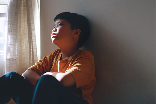 In the shadows of a home corner, a little Asian boy with a sad and lonely expression hides. Only his face is illuminated by the shadowy light coming through a window.