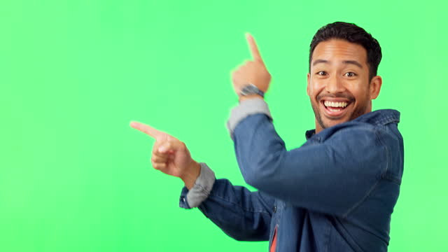 Asian man, pointing and dancing on green screen in product placement or advertisement against studio background. Portrait of happy male showing gesture or point for advertising or marketing on mockup
